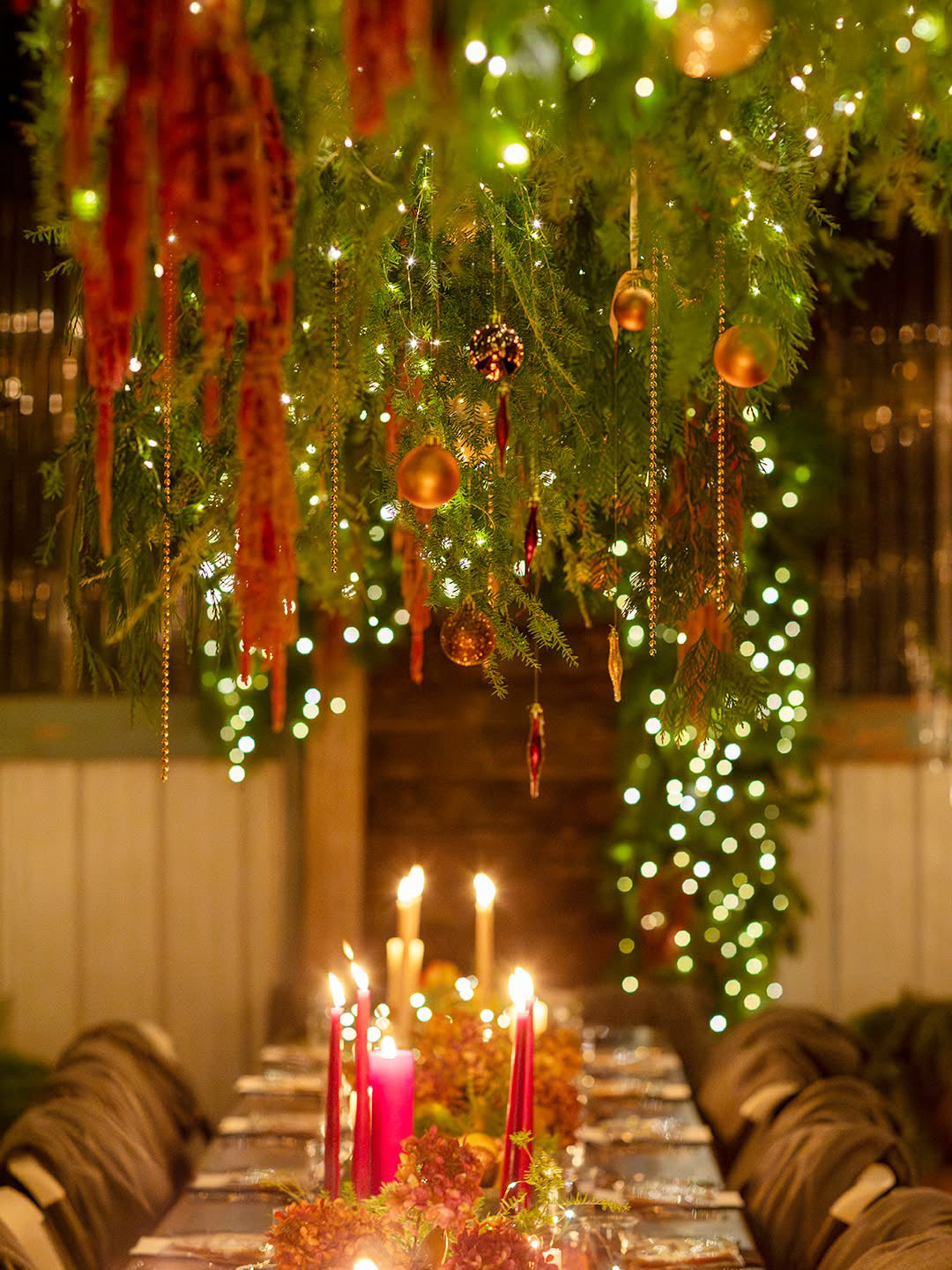 A festive holiday table setting under a lush green garland canopy adorned with warm white string lights, gold ornaments, and red dried floral accents. Lit red and gold taper candles sit on the table runner, with wrapped chairs on both sides.