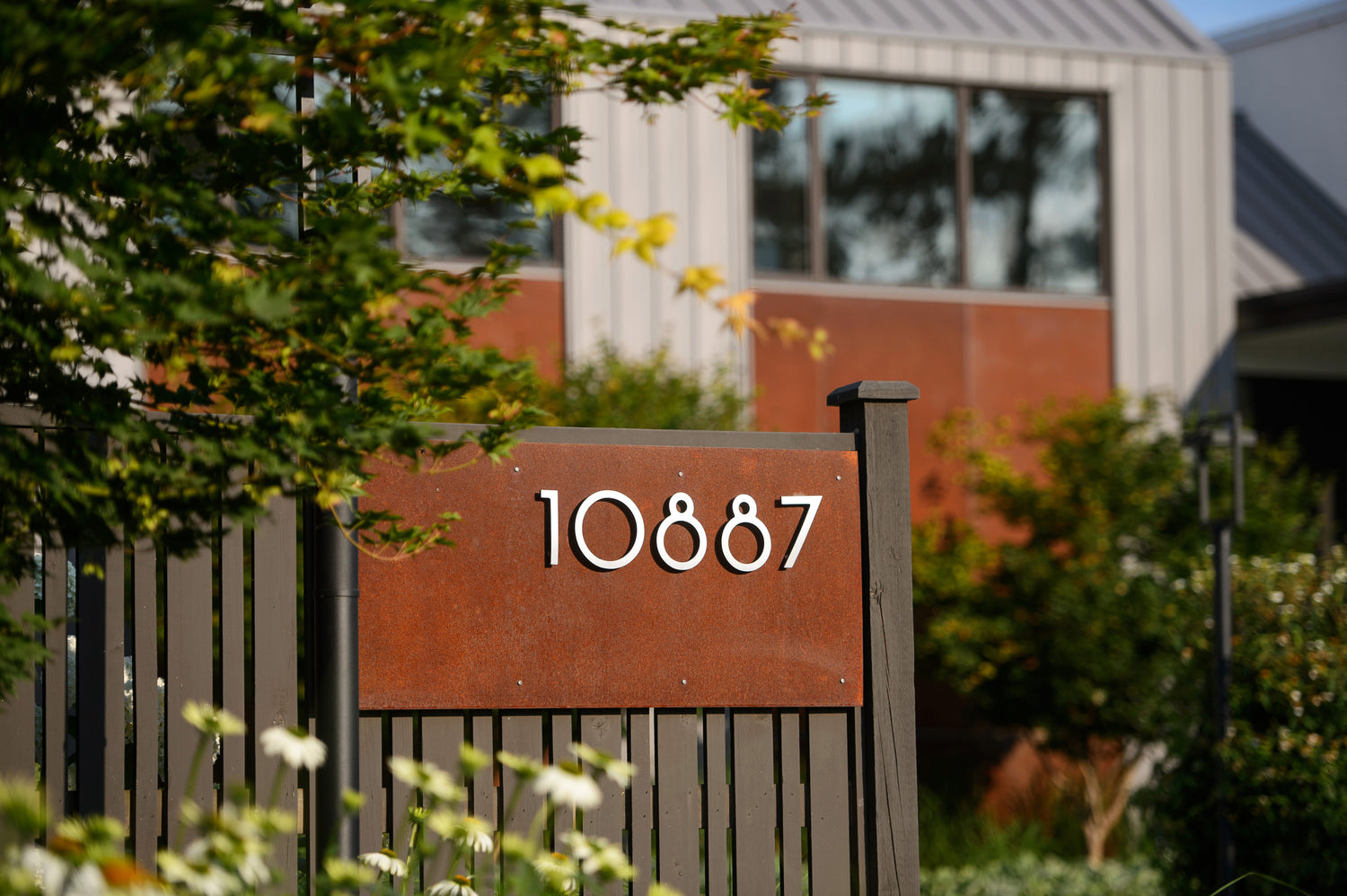 Close-up view of corten steel address sign on fence surrounded with blurred out greenery.  